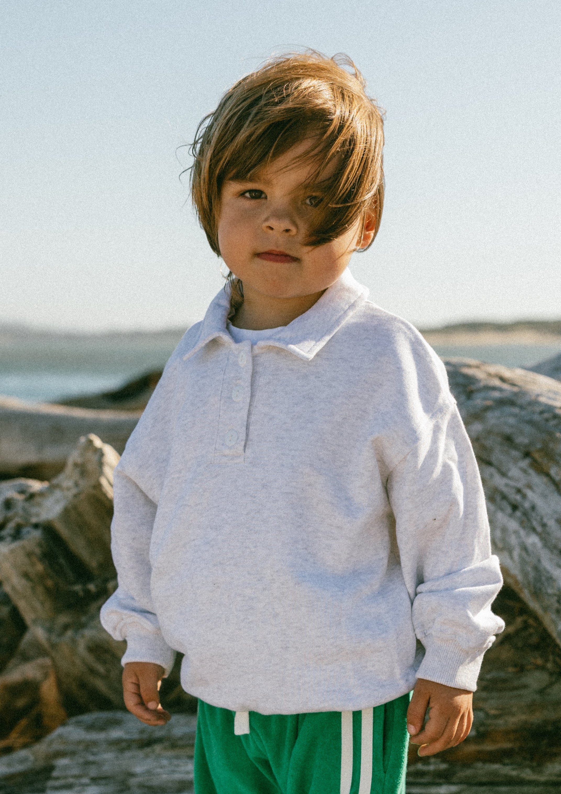Child standing in a natural setting with large logs and a clear sky.