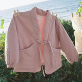 A dusty pink open-front jacket with a tie waist and large front pockets, hanging on a clothesline outdoors with the ocean and greenery in the background.