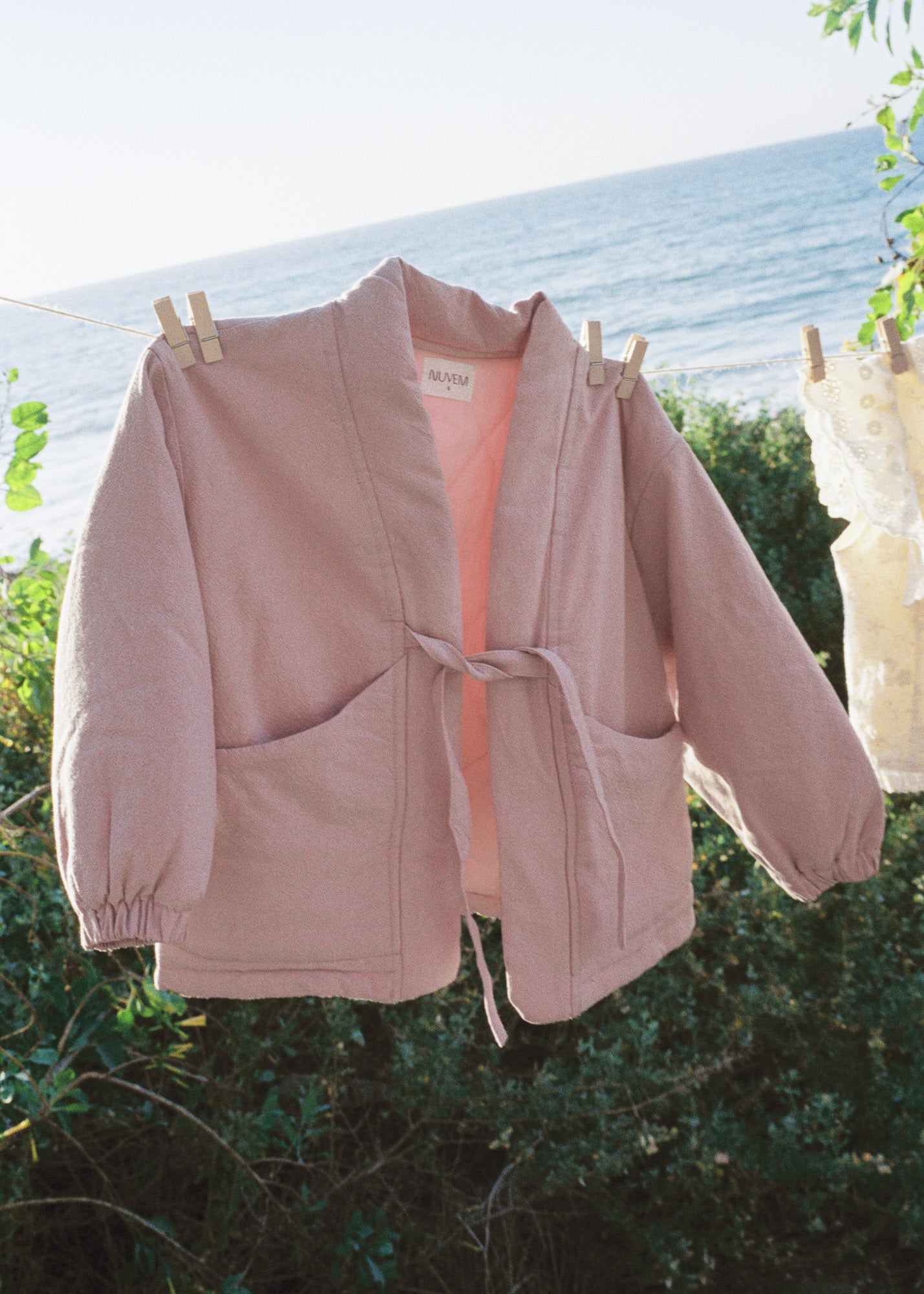 A dusty pink open-front jacket with a tie waist and large front pockets, hanging on a clothesline outdoors with the ocean and greenery in the background.