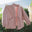 A dusty pink open-front jacket with a tie waist and large front pockets, hanging on a clothesline outdoors with the ocean and greenery in the background.