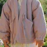 A child wearing a dusty pink hooded jacket with drawstrings and elastic cuffs, paired with white shorts, standing outdoors against a blurred green background.