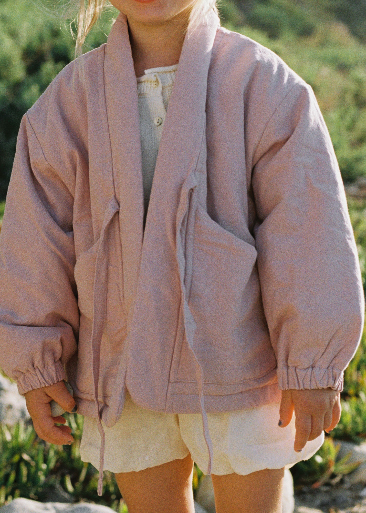 A child wearing a dusty pink hooded jacket with drawstrings and elastic cuffs, paired with white shorts, standing outdoors against a blurred green background.