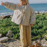A young girl stands on a sandy beach with green vegetation and the ocean in the background, wearing a cream quilted jacket and beige wide-leg pants. A woven straw bag is in the foreground.