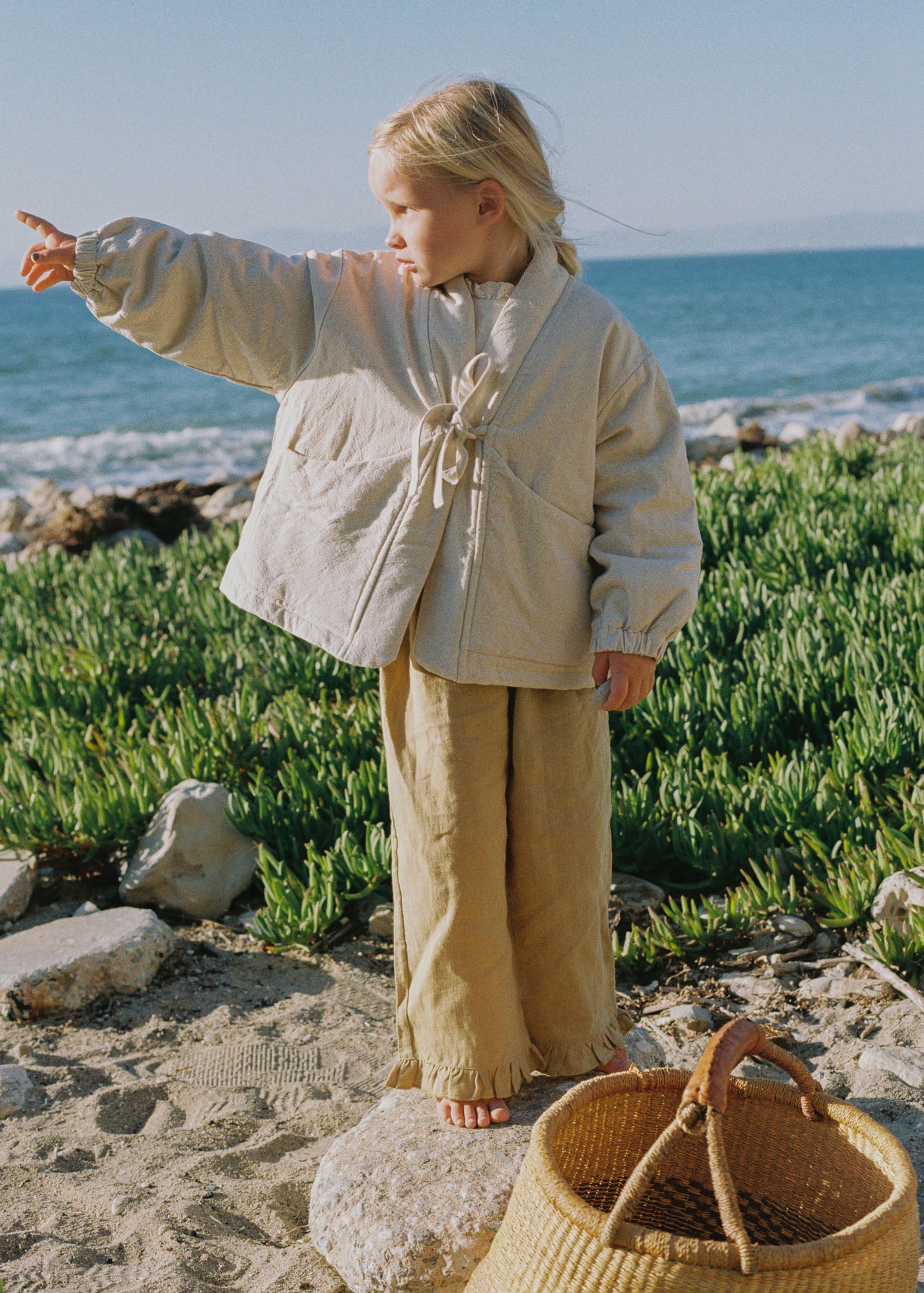A young girl stands on a sandy beach with green vegetation and the ocean in the background, wearing a cream quilted jacket and beige wide-leg pants. A woven straw bag is in the foreground.