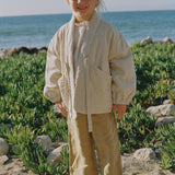 A young child standing on a sandy beach wearing a cream-colored linen jacket and matching wide-leg pants, with green beach plants and the ocean in the background.
