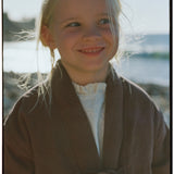 Child wearing the Amélie Coat in Cacao brown over a cream ruffled top at the beach