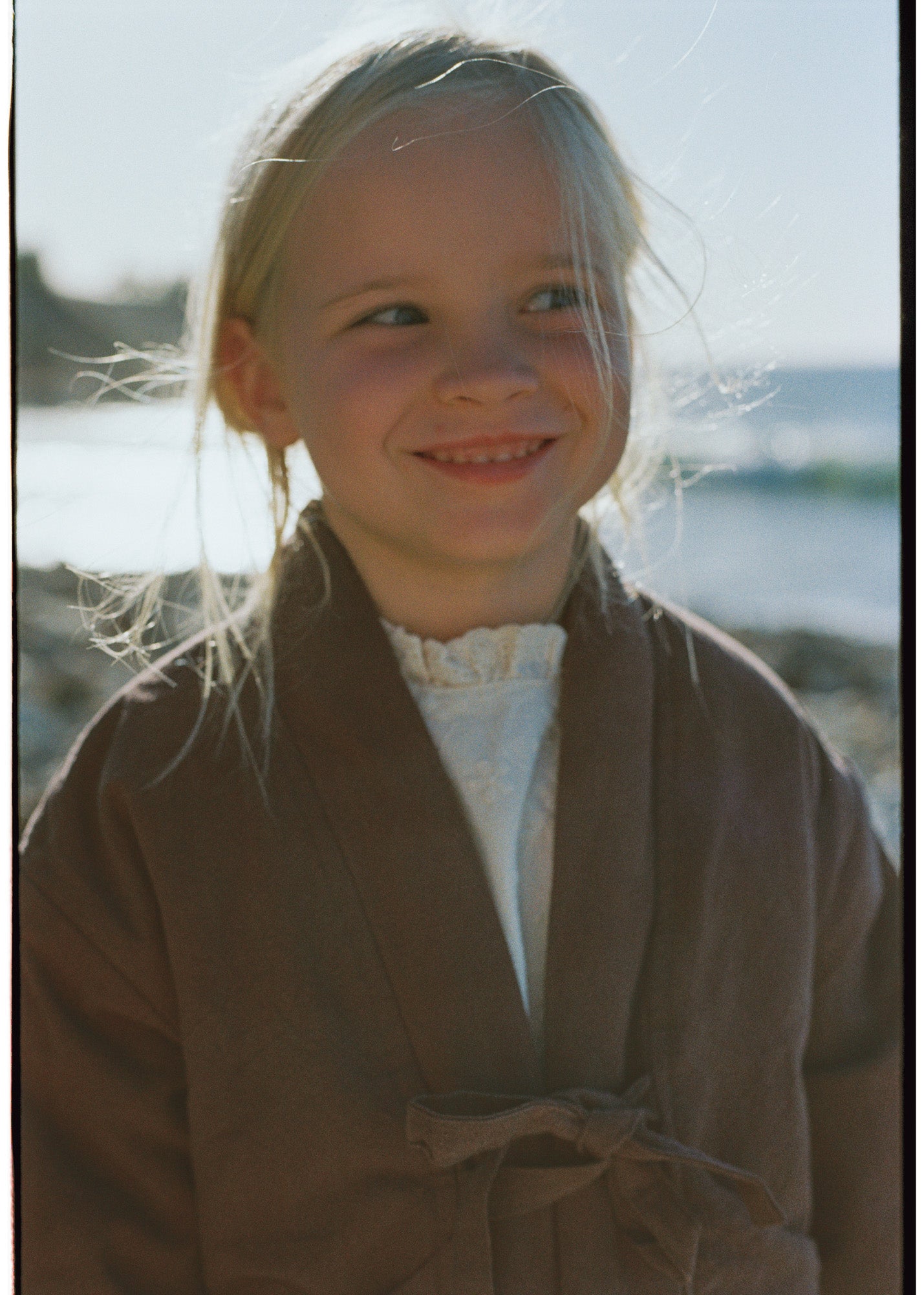 Child wearing the Amélie Coat in Cacao brown over a cream ruffled top at the beach