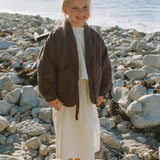 Full-length view of a child wearing the Amélie Coat in Cacao brown over a cream outfit on a rocky beach