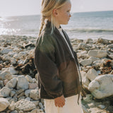 A young girl with blonde hair in a side braid stands on a rocky beach, wearing a dark olive green lightweight zip-up jacket over a white top and white skirt, looking towards the ocean.