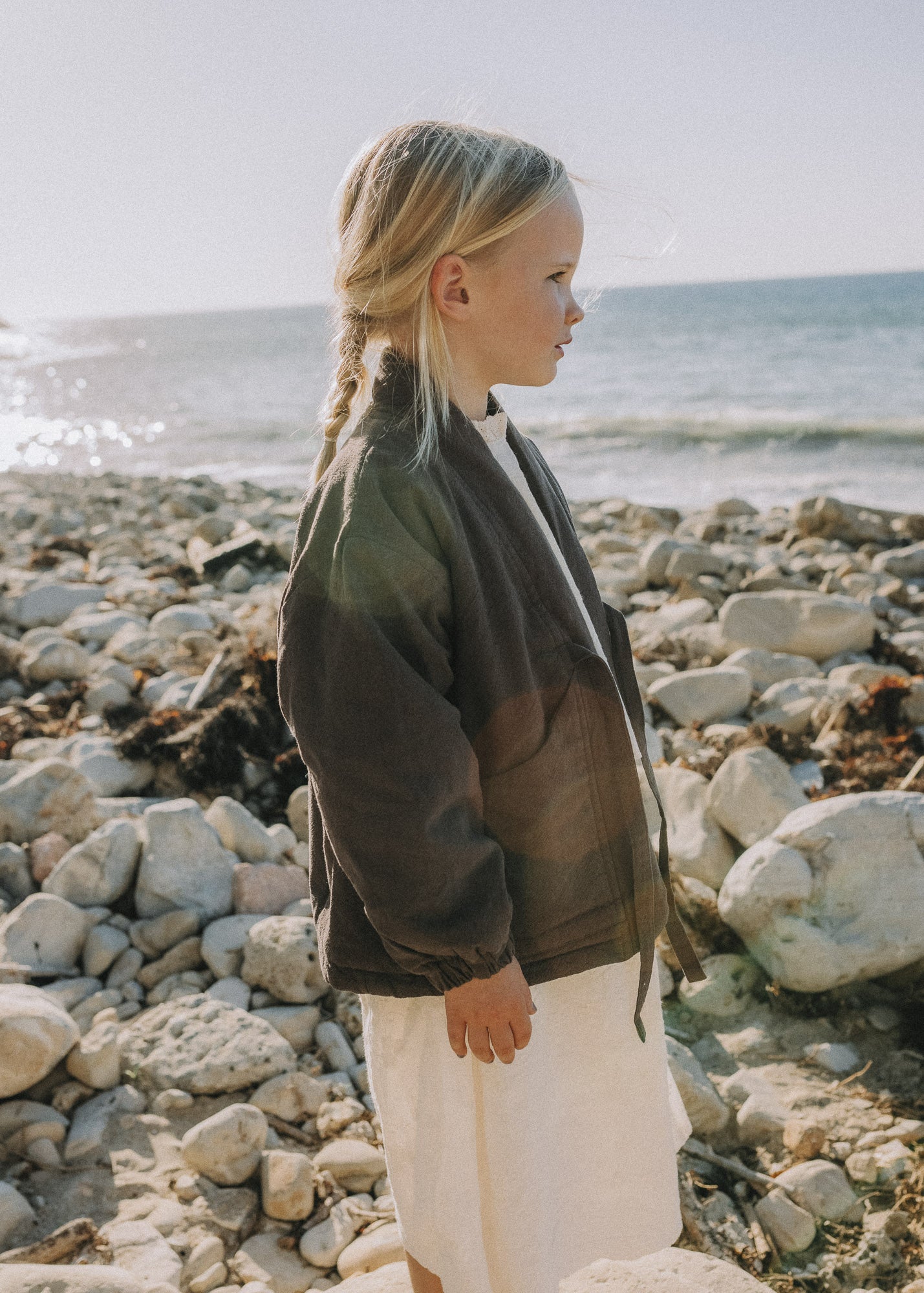 A young girl with blonde hair in a side braid stands on a rocky beach, wearing a dark olive green lightweight zip-up jacket over a white top and white skirt, looking towards the ocean.