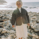 A young child stands barefoot on a large rock on a rocky beach, wearing a dark olive green wrap-style jacket over a white top and a long white skirt, with the ocean in the background.