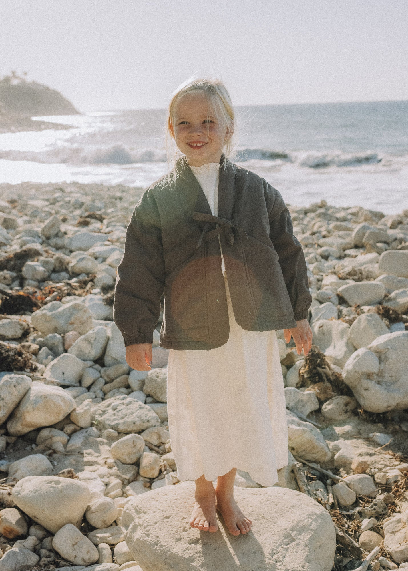 A young child stands barefoot on a large rock on a rocky beach, wearing a dark olive green wrap-style jacket over a white top and a long white skirt, with the ocean in the background.