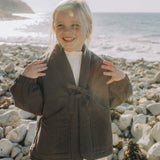 A young blonde girl smiling on a rocky beach, wearing a dark brown belted jacket over a cream dress.