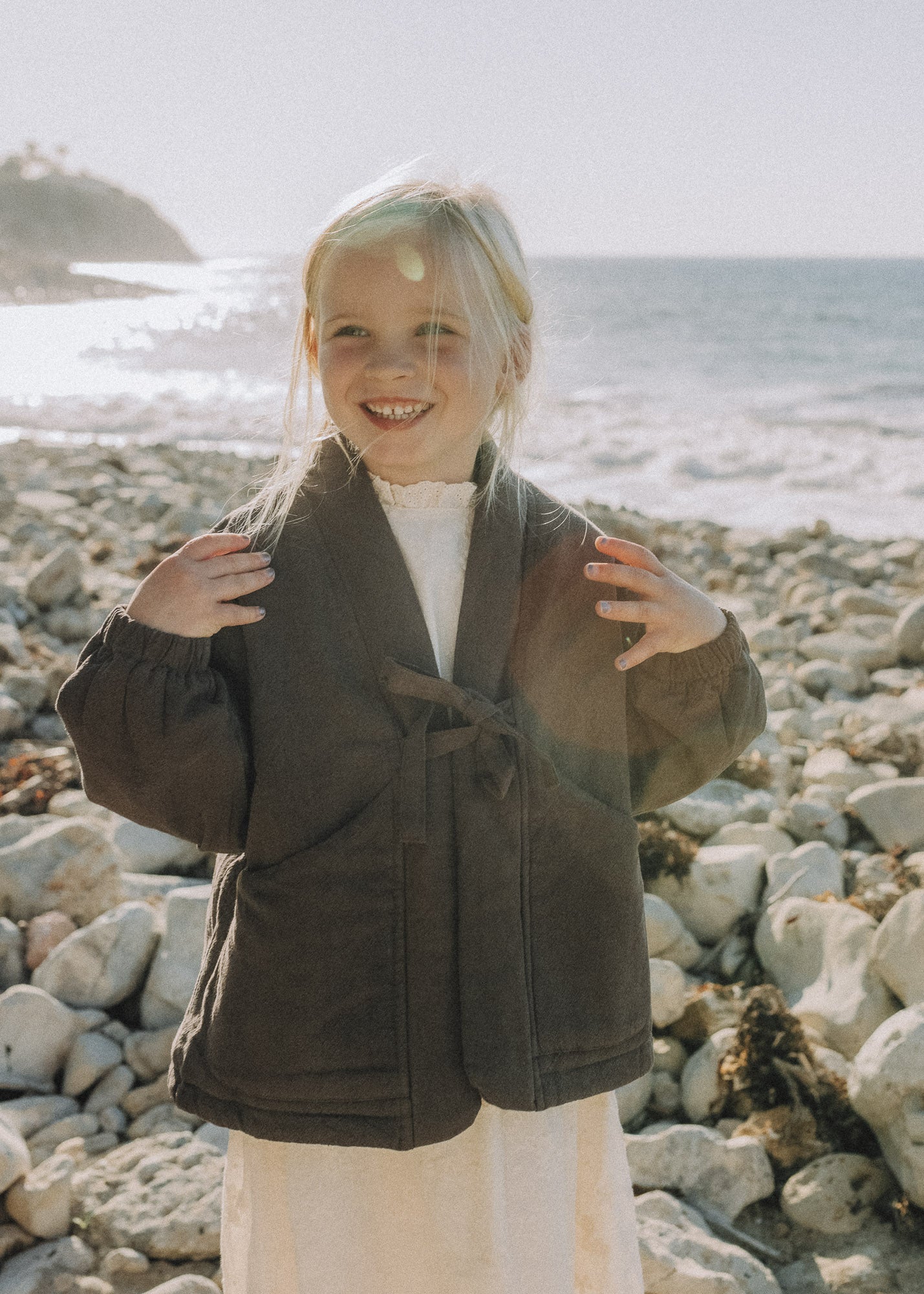 A young blonde girl smiling on a rocky beach, wearing a dark brown belted jacket over a cream dress.