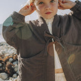A young girl with blonde hair stands on a rocky beach wearing a dark brown linen jacket with a tie closure over a white top and skirt.