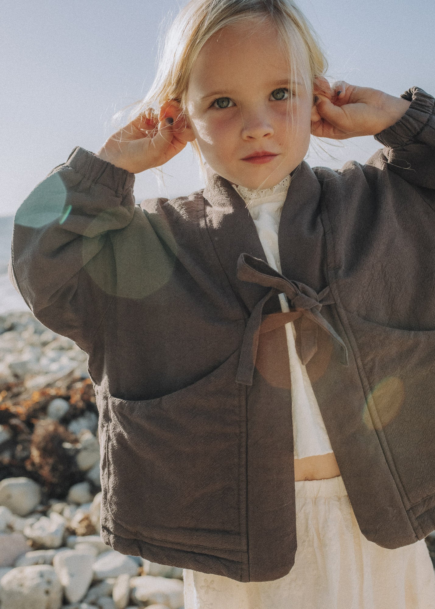 A young girl with blonde hair stands on a rocky beach wearing a dark brown linen jacket with a tie closure over a white top and skirt.