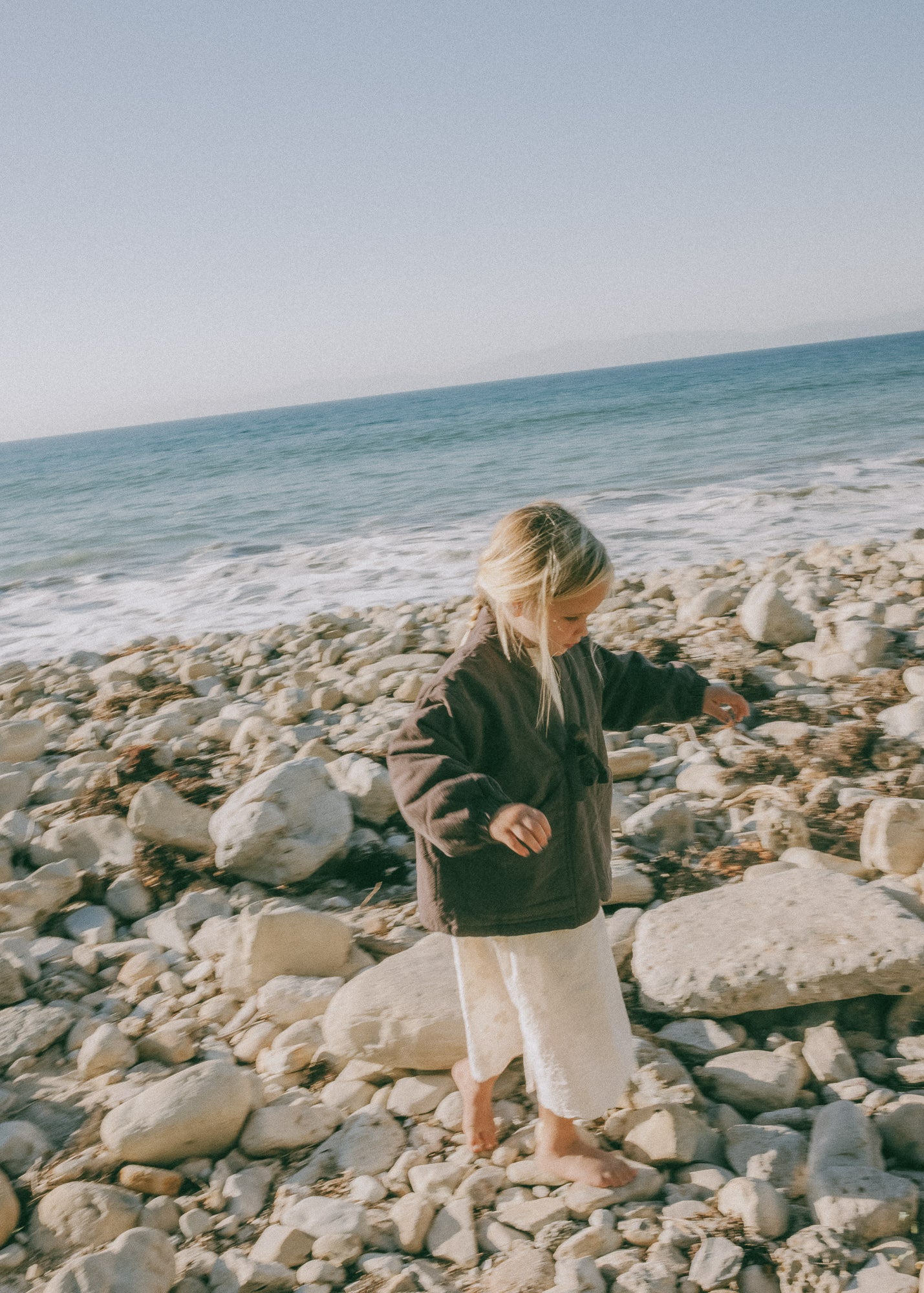 Child walking across a rocky beach wearing the Amélie Coat in Cacao brown over a cream outfi