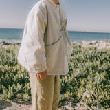 A young child standing outdoors on a sandy beach wearing a cream-colored linen jacket and beige trousers, with the ocean and green vegetation in the background.
