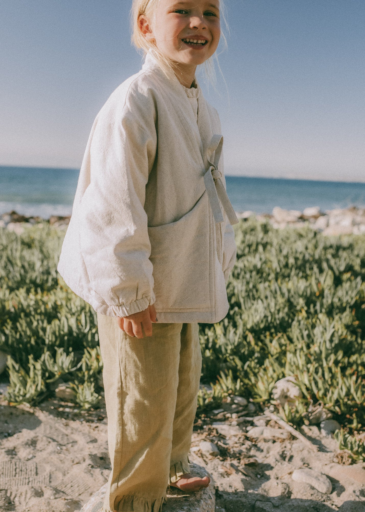 A young child standing outdoors on a sandy beach wearing a cream-colored linen jacket and beige trousers, with the ocean and green vegetation in the background.