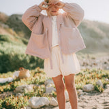 A young child standing outdoors on a sandy, rocky beach wearing a dusty pink quilted jacket over a white top and white shorts.