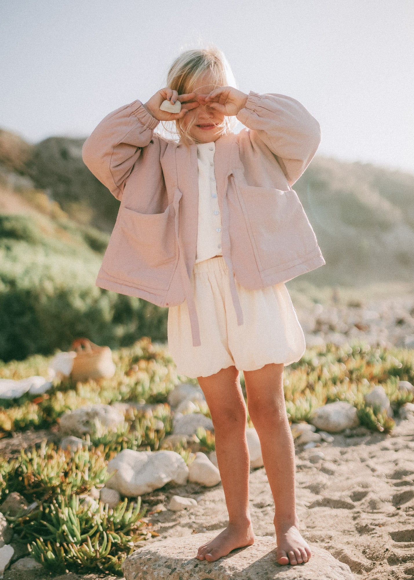 A young child standing outdoors on a sandy, rocky beach wearing a dusty pink quilted jacket over a white top and white shorts.