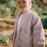 A young blonde girl smiling outdoors, wearing a light dusty pink linen-blend jacket over a white top, with a blurred natural hillside background.