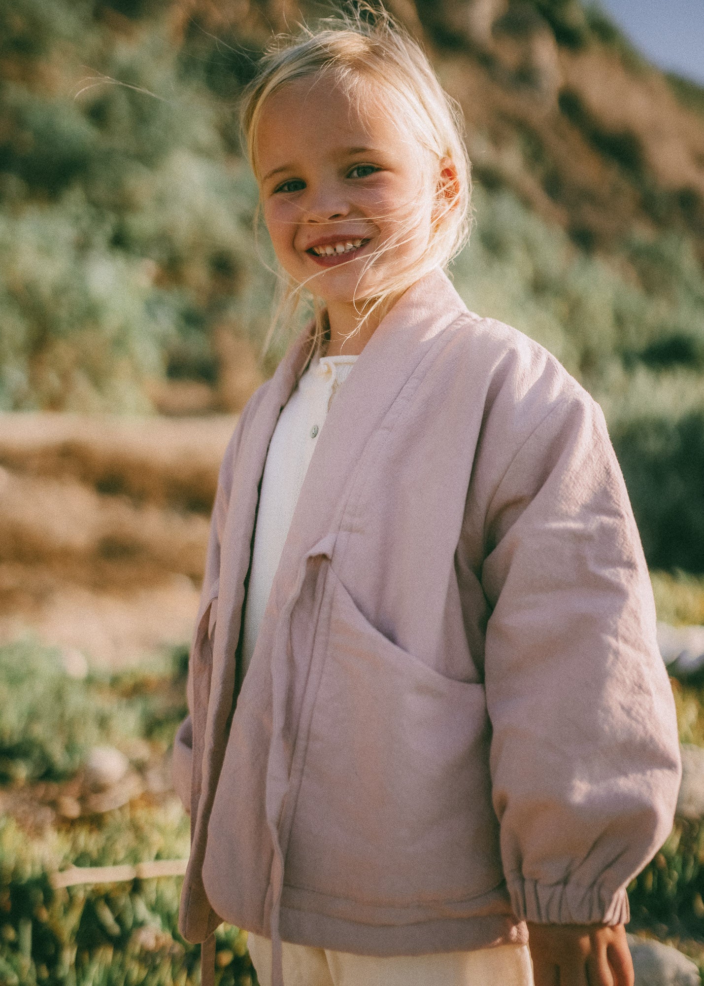 A young blonde girl smiling outdoors, wearing a light dusty pink linen-blend jacket over a white top, with a blurred natural hillside background.