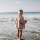 A young girl with blonde hair in a braid stands on a sandy beach at the water's edge, viewed from behind. She is wearing a light pink jacket and white shorts, looking out towards the ocean waves under a clear blue sky.