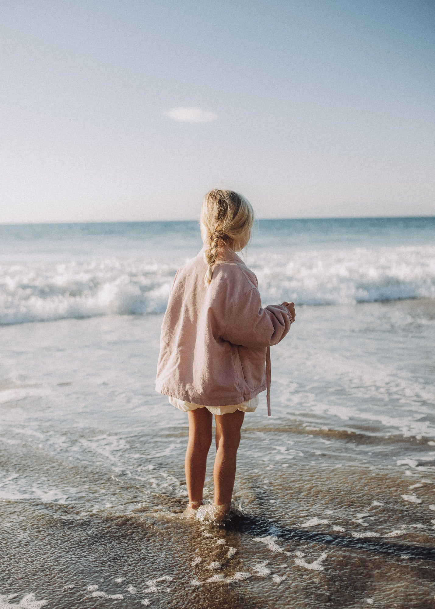 A young girl with blonde hair in a braid stands on a sandy beach at the water's edge, viewed from behind. She is wearing a light pink jacket and white shorts, looking out towards the ocean waves under a clear blue sky.