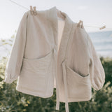 A light beige linen jacket with large front patch pockets hangs on a clothesline outdoors with a blurred beach and foliage background.