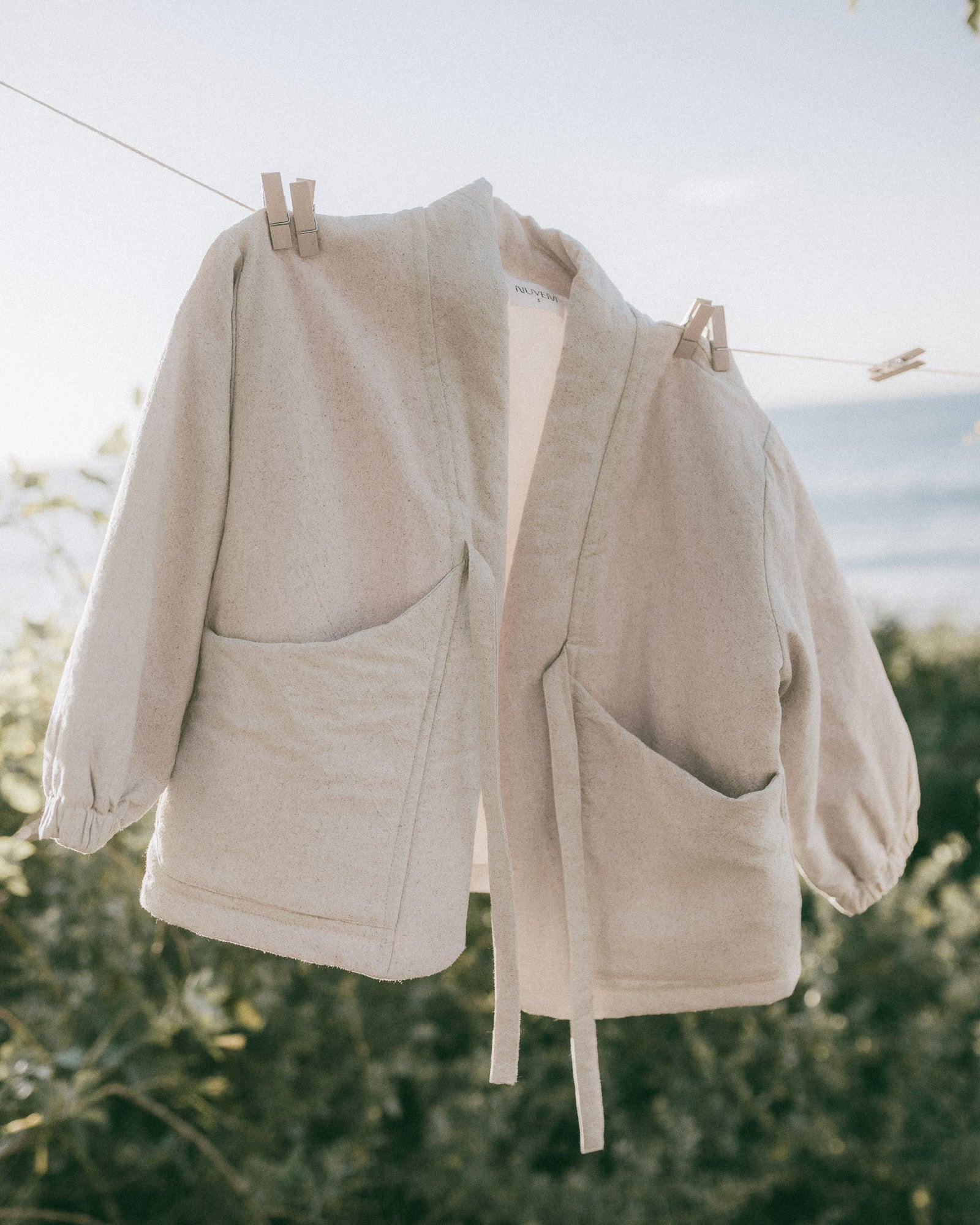A light beige linen jacket with large front patch pockets hangs on a clothesline outdoors with a blurred beach and foliage background.