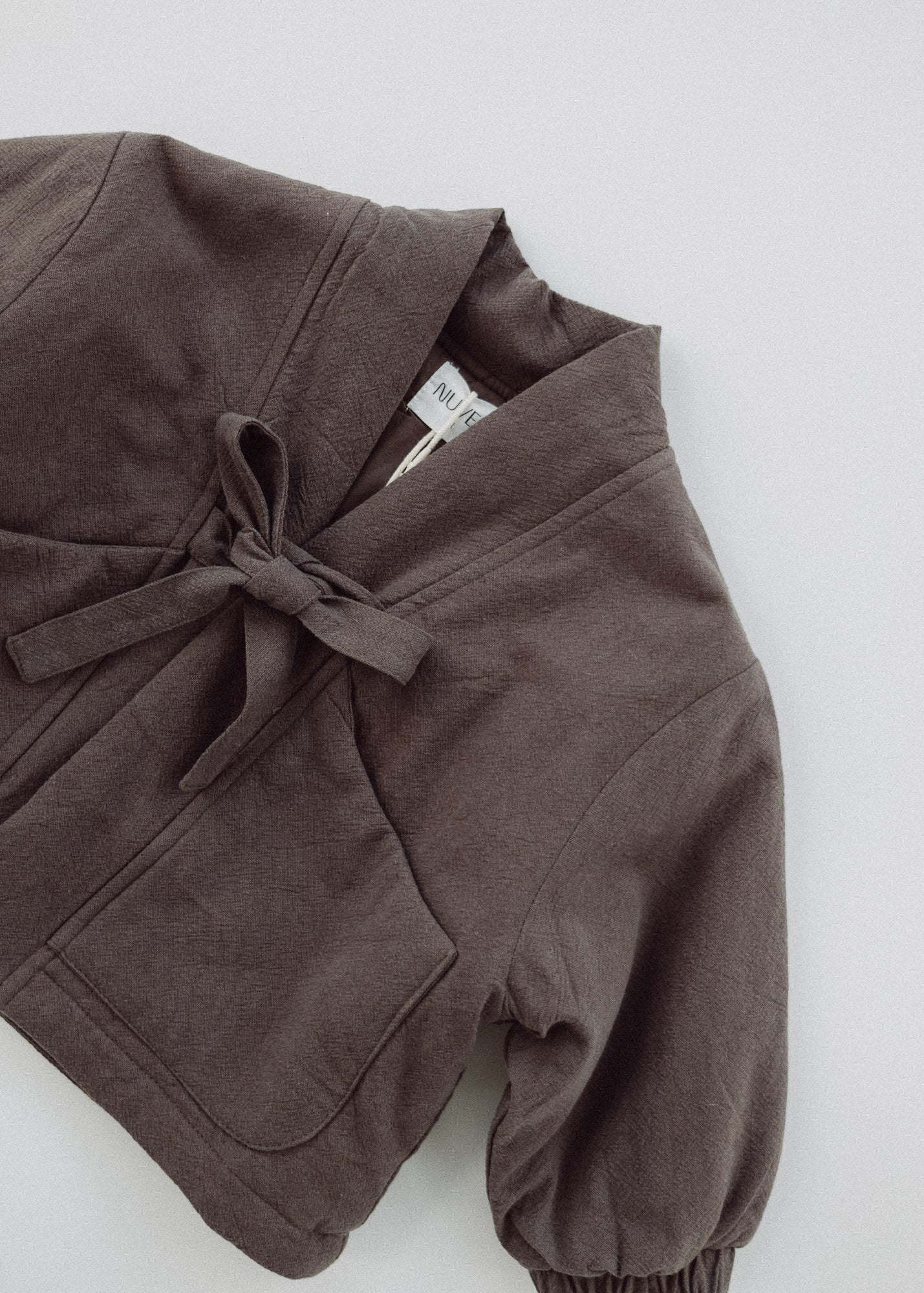 Flat-lay detail of the Amélie Coat in Cacao brown, showing the fabric texture, front bow and patch pocket