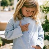 A young girl with blonde hair looking down while wearing a light blue knit cardigan with white buttons and a white skirt, standing outdoors in a garden setting with white flowers.