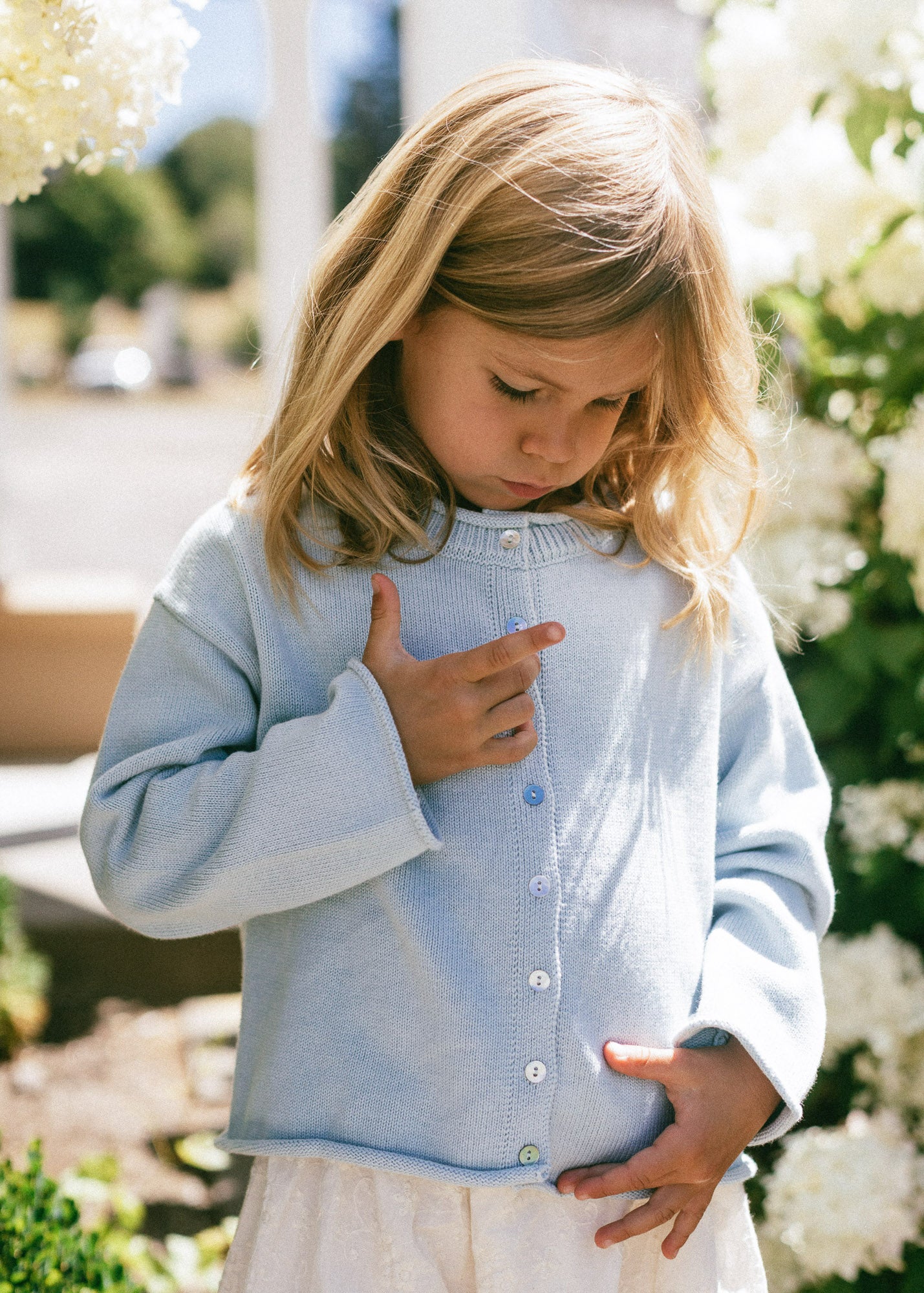 A young girl with blonde hair looking down while wearing a light blue knit cardigan with white buttons and a white skirt, standing outdoors in a garden setting with white flowers.