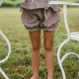 A child wearing a matching ruffled linen romper in a muted dusty mauve color, standing barefoot on grass next to a white metal garden table and chair.