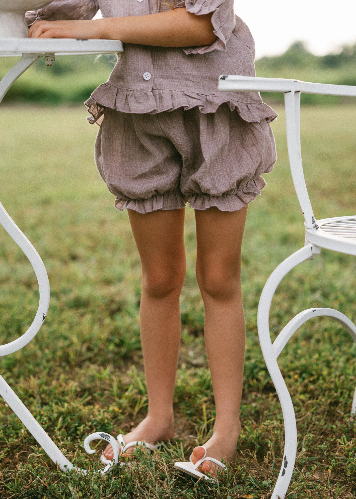 A child wearing a matching ruffled linen romper in a muted dusty mauve color, standing barefoot on grass next to a white metal garden table and chair.