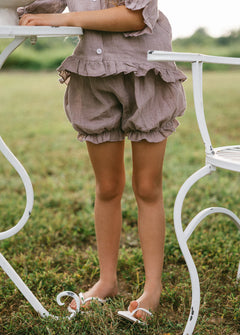 A child wearing a matching ruffled linen romper in a muted dusty mauve color, standing barefoot on grass next to a white metal garden table and chair.
