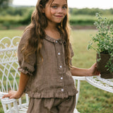 A young girl with long brown hair stands outdoors in a grassy yard next to a white metal table, wearing a matching brown linen-look two-piece outfit consisting of a button-front top and ruffled shorts.