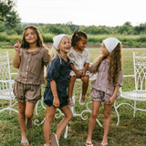Four young girls wearing matching two-piece linen outfits and white headbands are posing together outdoors on a grassy lawn with white garden chairs and trees in the background.