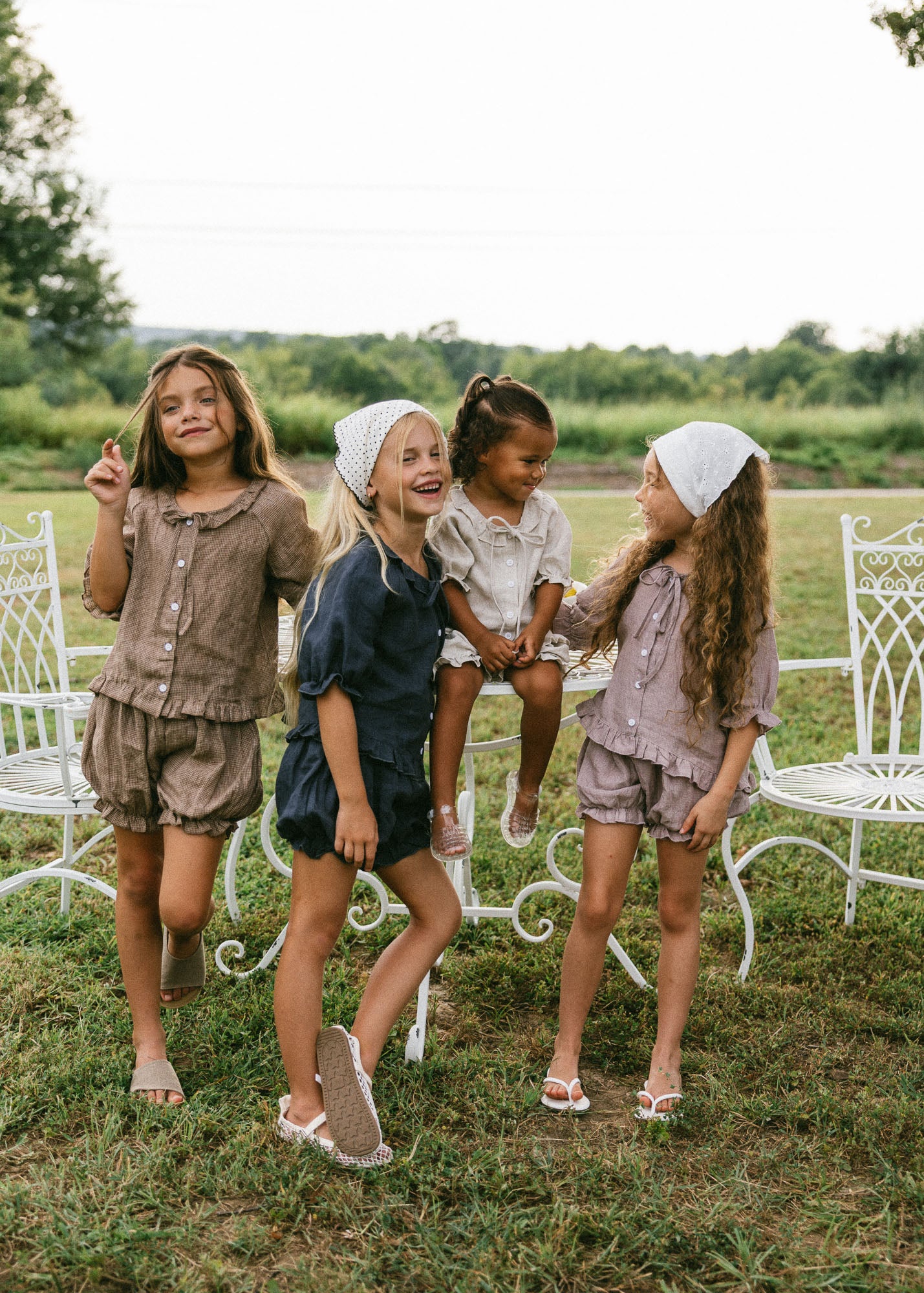 Four young girls wearing matching two-piece linen outfits and white headbands are posing together outdoors on a grassy lawn with white garden chairs and trees in the background.