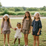 Four young girls standing together on a grassy field outdoors, wearing matching linen-like clothing sets in muted brown, taupe, navy, and white. The background shows a blurred green landscape and trees under a bright sky.